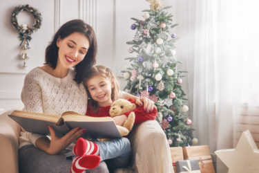 Mom reading a book to her cute daughter near Christmas tree indoors.