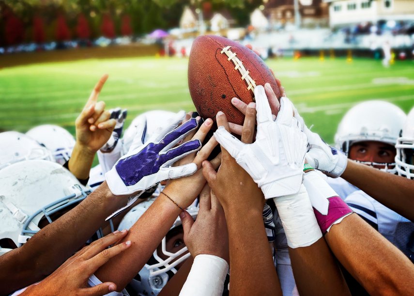 football players huddles in a circle reaching for the football above